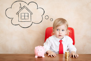 Toddler in a shirt and tie sitting at a desk with a piggy bank and a stack of coins, with a thought bubble showing a drawing of a house